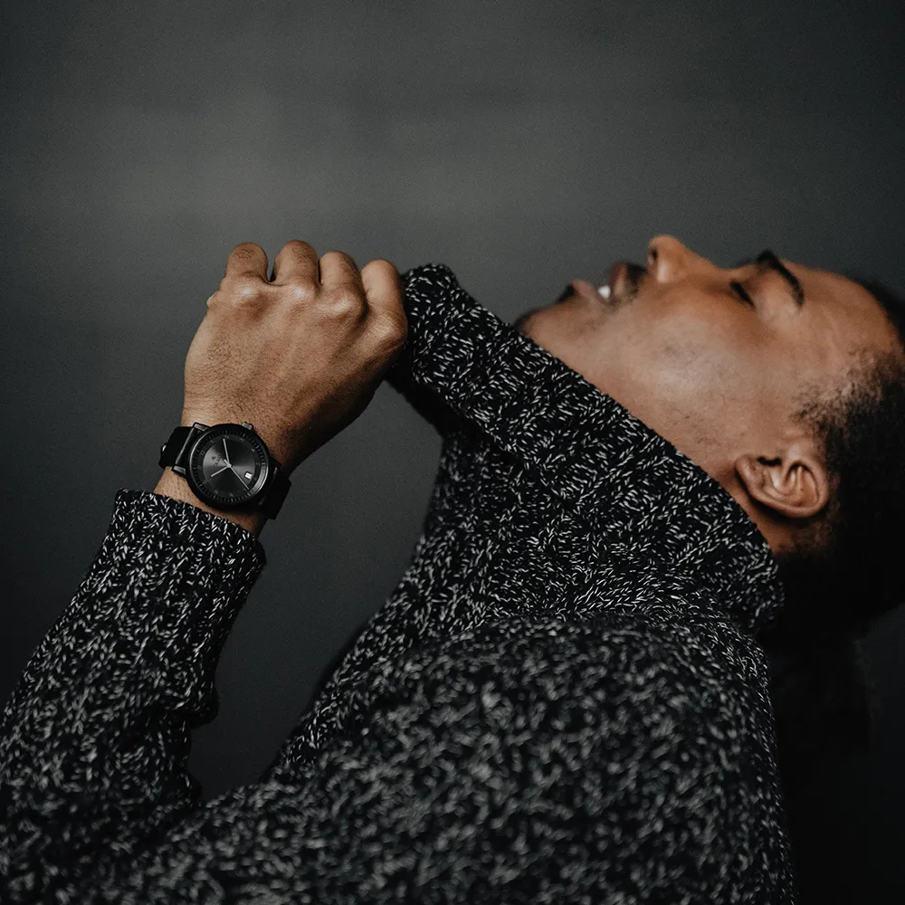 Head tipped back, a hand at the collar of a marled knit, a black Aspect on the wrist